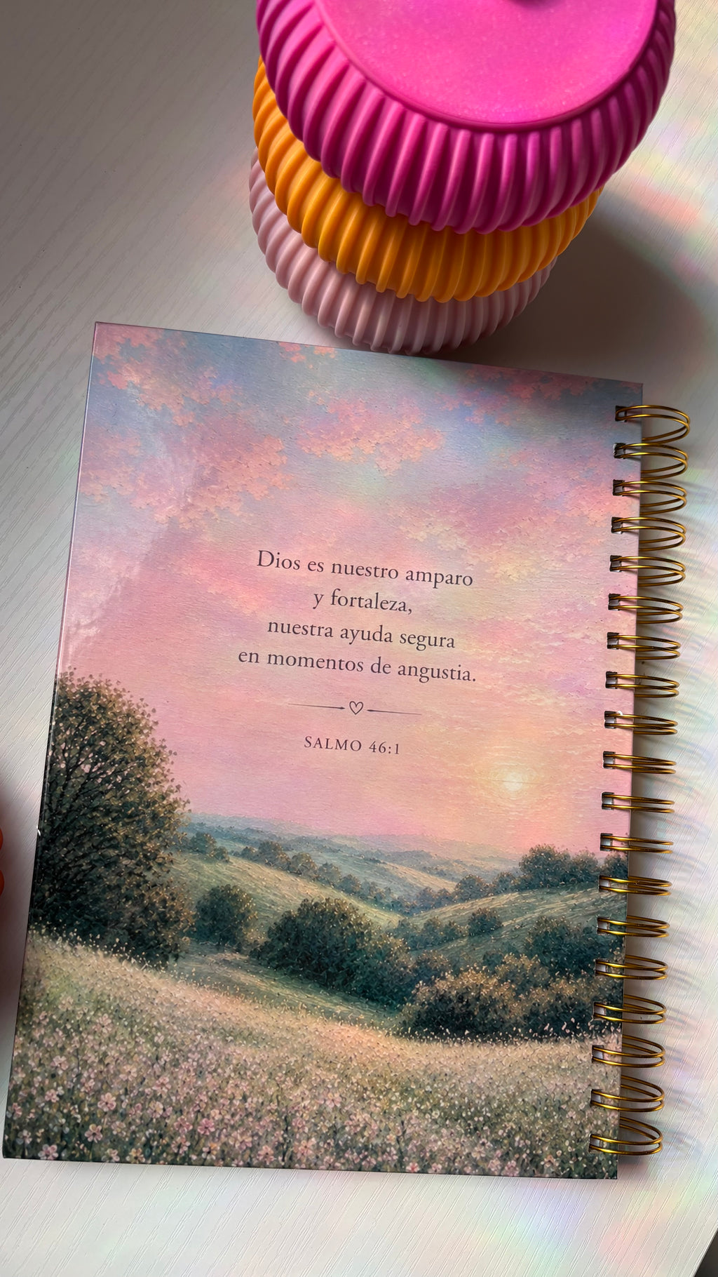 Notebook with a scenic cover and text, placed on a surface with a pink and yellow container.
