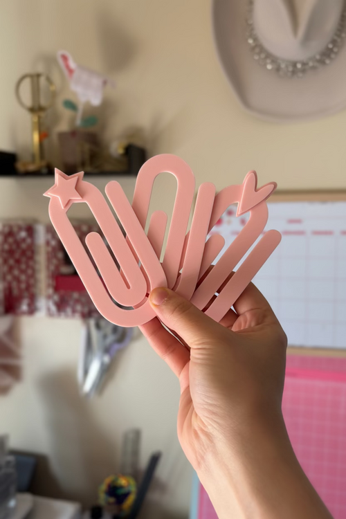Hand holding a pink paper clip shaped like a peacock in a room with various items on a shelf.